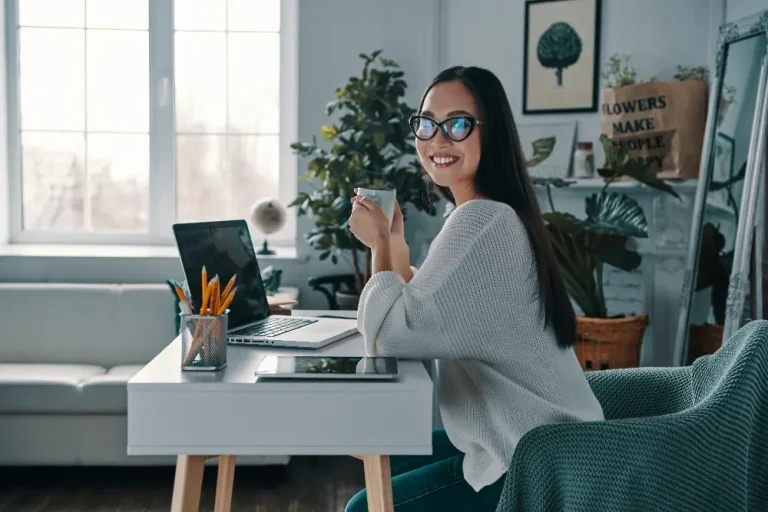 A woman wearing glasses sits at a desk, focused on her laptop while working.